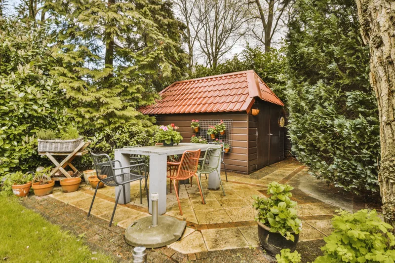 Cozy backyard garden with a wooden shed, terracotta roof, and a patio area featuring a weathered white table with colorful chairs surrounded by lush greenery.